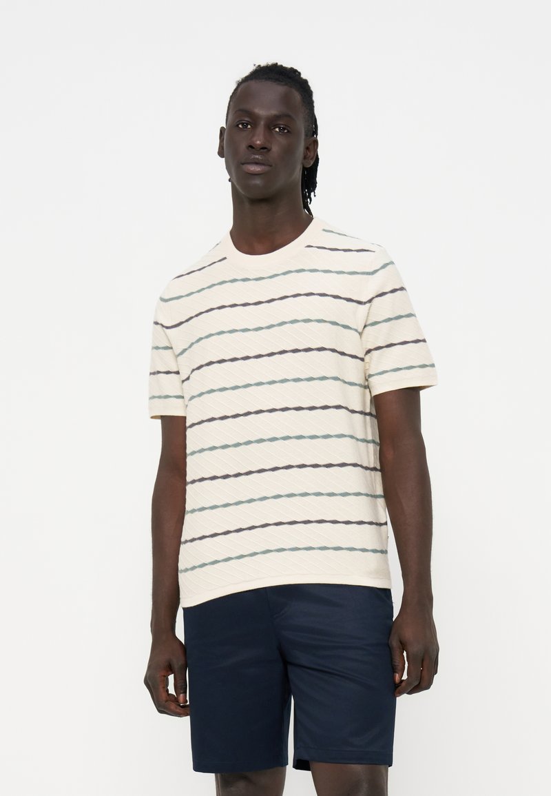 Junger Mann mit dunkler Haut und Dreadlocks trägt ein cremefarbenes kurzärmeliges Hemd mit dünnen horizontalen Streifen und dunkelblaue Shorts.
