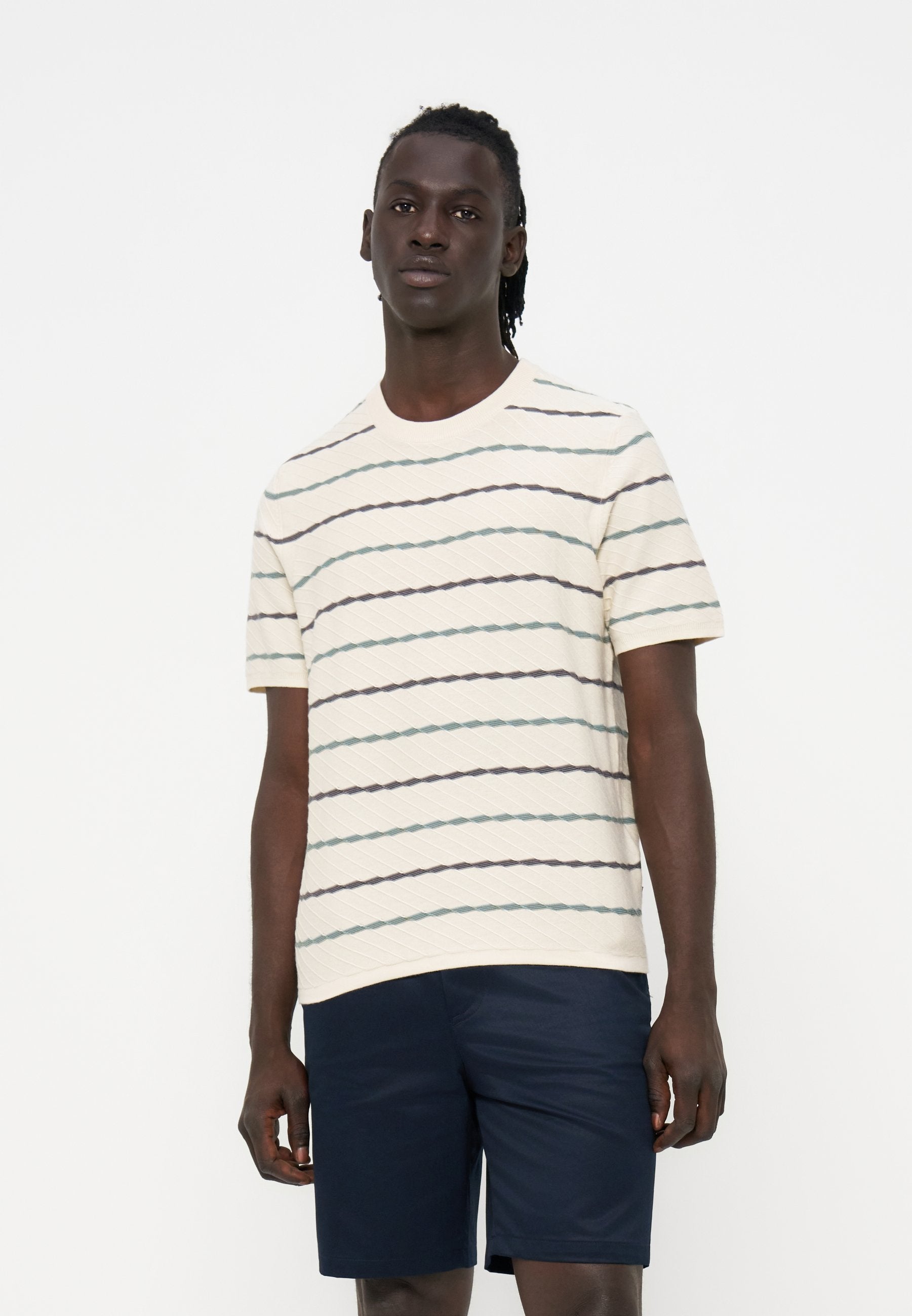Junger Mann mit dunkler Haut und Dreadlocks trägt ein cremefarbenes kurzärmeliges Hemd mit dünnen horizontalen Streifen und dunkelblaue Shorts.