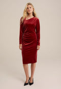 Rotes Samtkleid mit langen Ärmeln, asymmetrischem Ausschnitt, figurbetontem Schnitt, Raffung an der Seite und knielangem Saum. Schwarze High Heels.