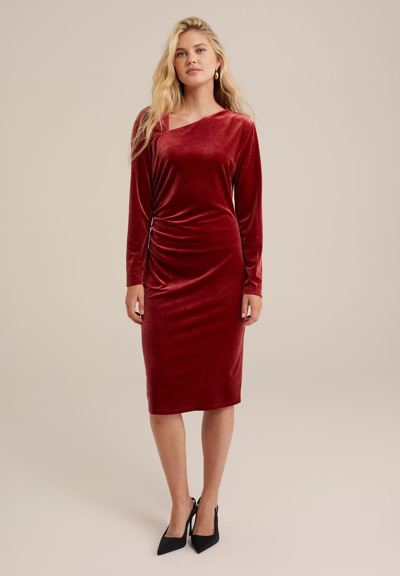 Rotes Samtkleid mit langen Ärmeln, asymmetrischem Ausschnitt, figurbetontem Schnitt, Raffung an der Seite und knielangem Saum. Schwarze High Heels.