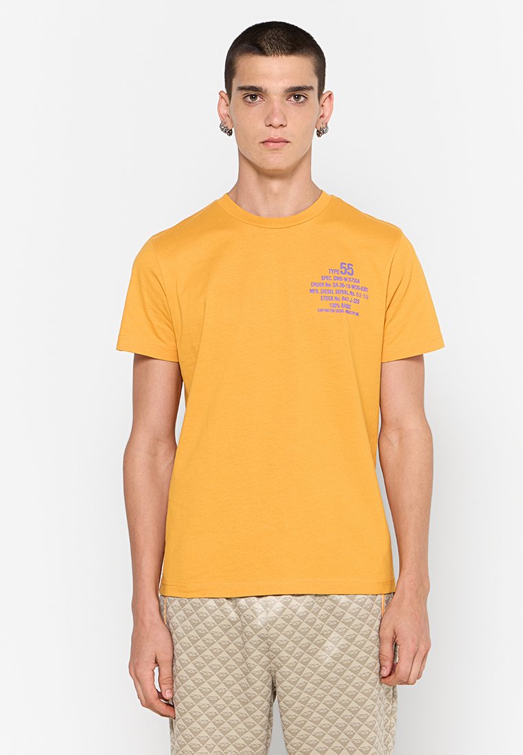 Gelbes Kurzarm-T-Shirt aus Baumwolle mit einem lila grafischen Druck auf der linken Brust, kombiniert mit strukturierten hellen Hosen.