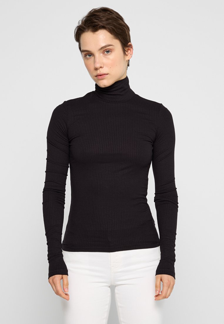 Junge Person mit kurzem Haar, die einen enganliegenden schwarzen gerippten Rollkragenpullover und weiße Hosen trägt, frontal vor einem schlichten Hintergrund.