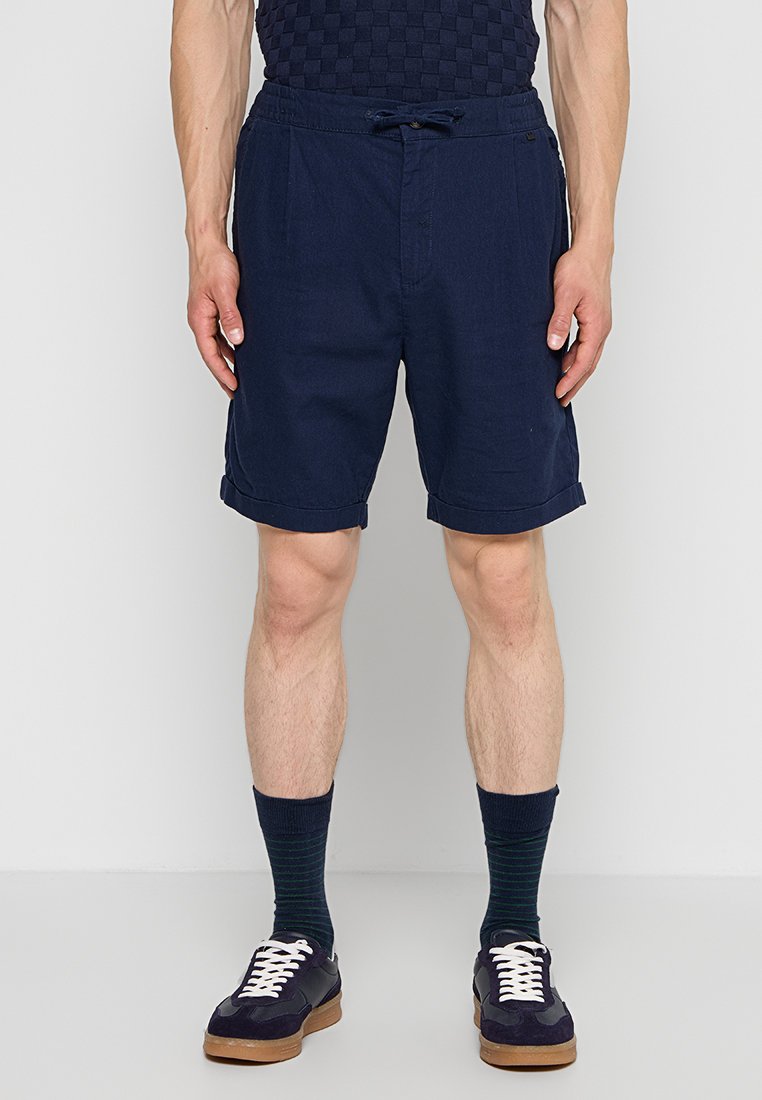 Mann trägt dunkelblaue Shorts mit umgeschlagenem Saum, dunkle gemusterte Socken und schwarze Sneakers mit weißen Schnürsenkeln vor einfarbigem Hintergrund.