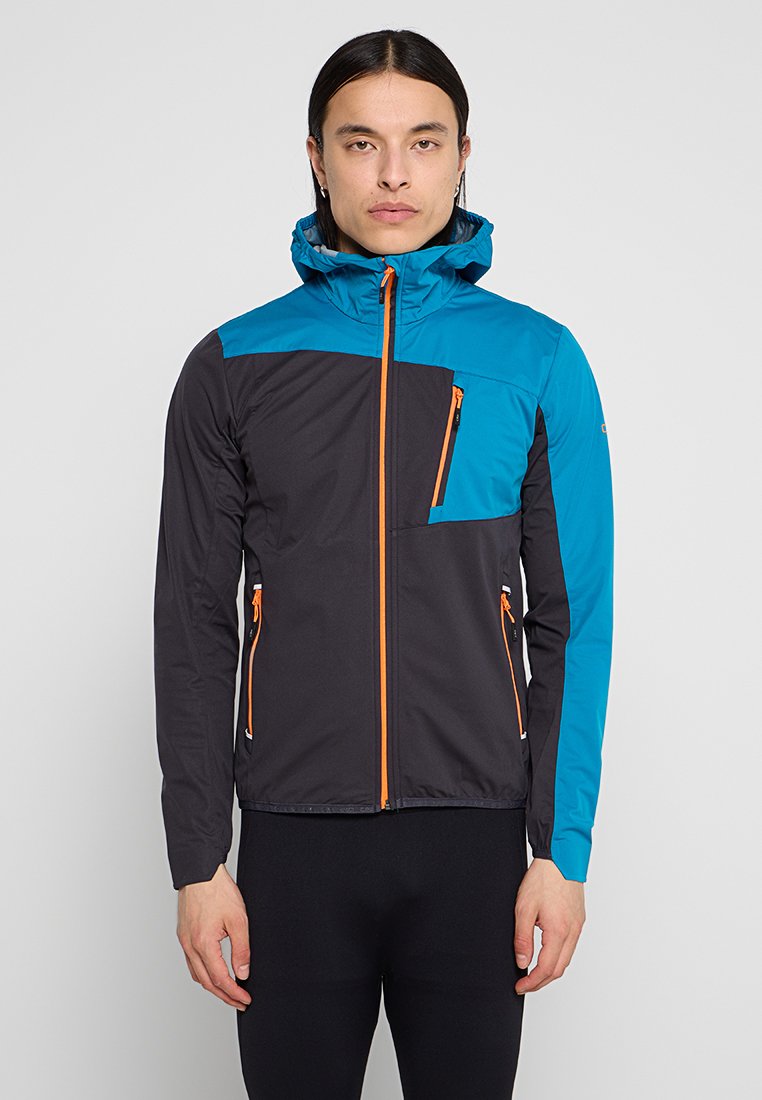 Mann in einer blau-schwarzen Kapuzenjacke mit orangefarbenen Reißverschlüssen, der vor einem neutralen grauen Hintergrund steht.