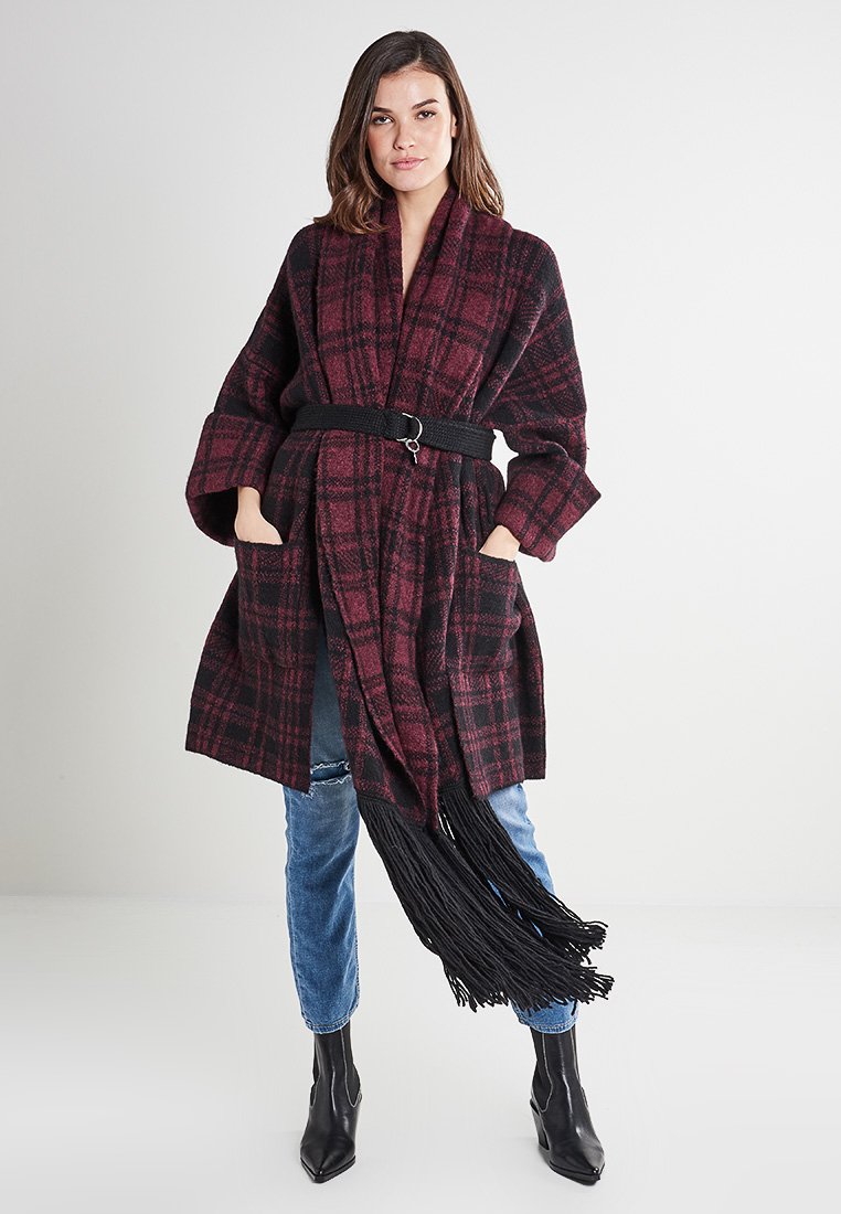 Karierte bordeaux-rote und schwarze Jacke mit Gürtel, langen Ärmeln, Taschen und fransigem Saum, aus einem strukturierten Material gefertigt.