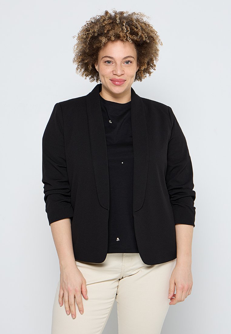 Schwarzer, strukturierter Blazer mit glatter Textur, offene Vorderseite und dreiviertellangen Ärmeln, getragen über einem schwarzen Hemd und hellen Hosen.