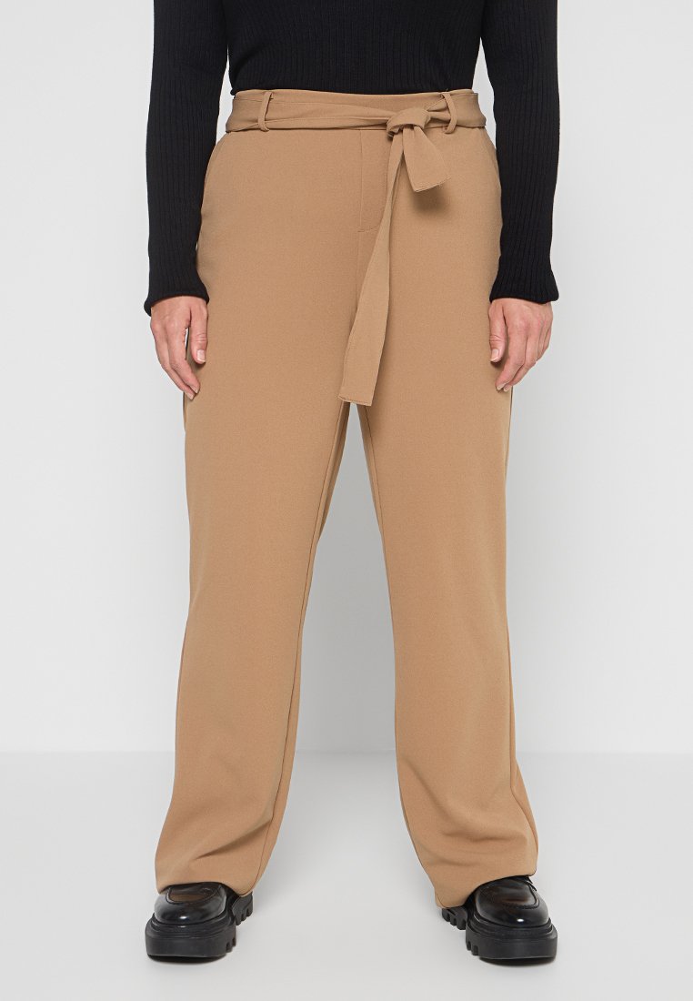 Helle beigefarbene Hochbundhose mit einem Stoffgürtel, weit geschnitten, und einer glatten Textur, kombiniert mit einem schwarzen, gerippten Langarmoberteil und klobigen schwarzen Schuhen.
