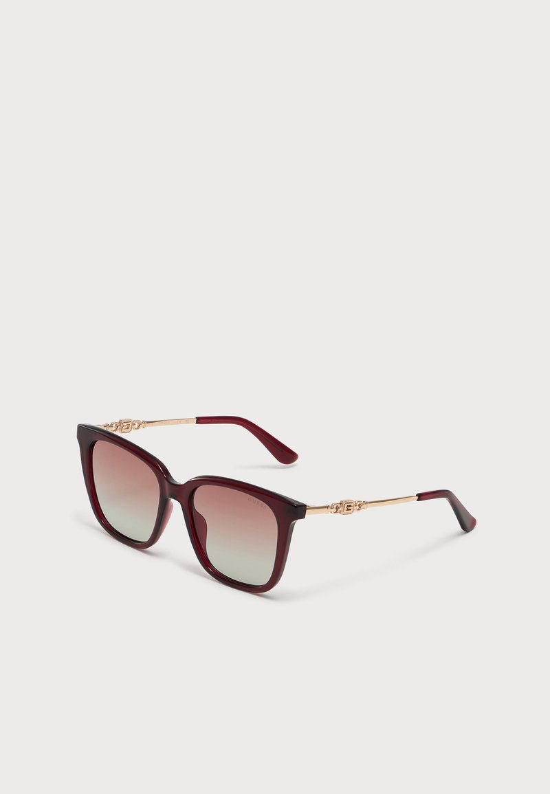 Quadratische Sonnenbrille mit dunkelrotem Rahmen, goldenen Bügeln und Verlaufsgläsern, auf einem schlichten hellen Hintergrund platziert.