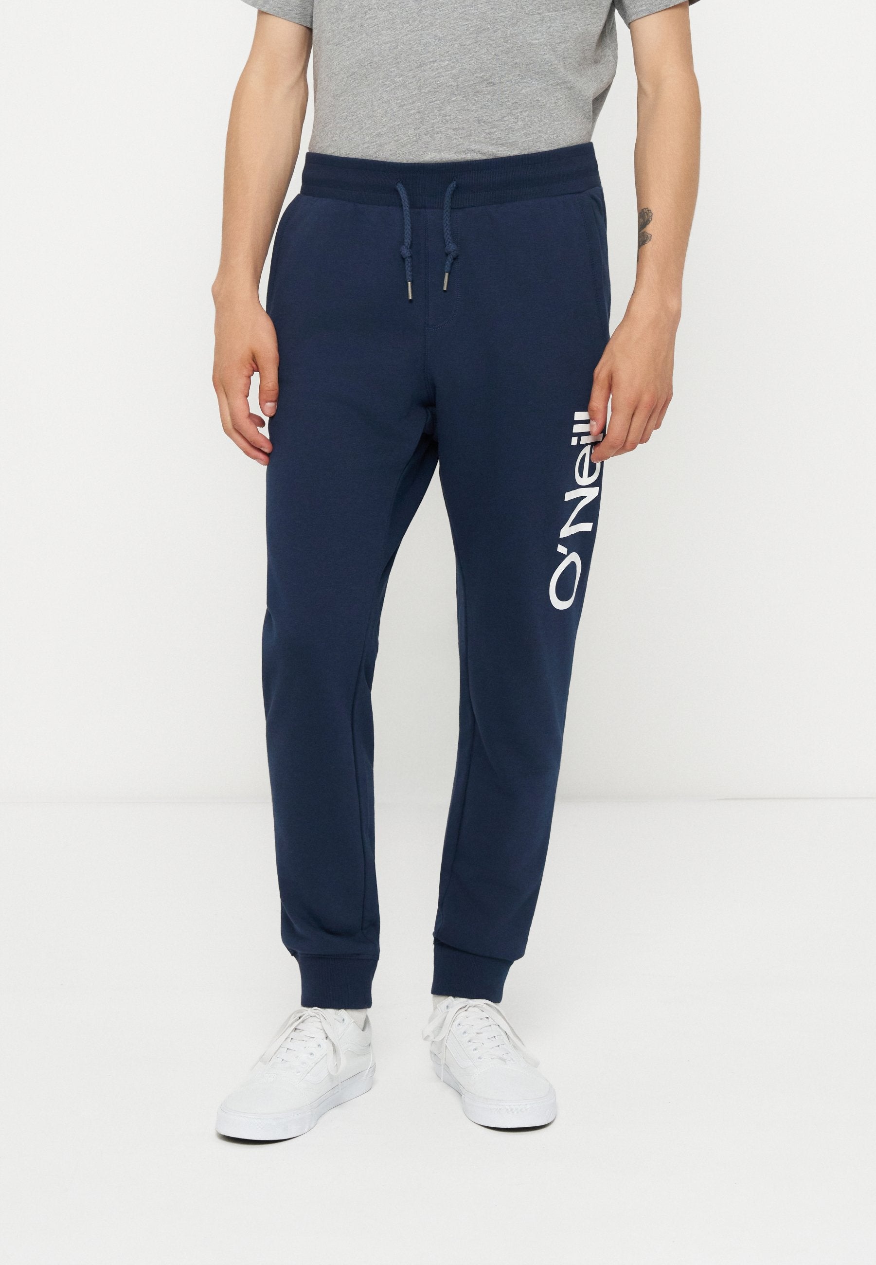 Mann trägt marineblaue Jogginghose mit O'Neill-Logo am linken Oberschenkel, graues T-Shirt und weiße Sneaker, steht vor einem schlichten weißen Hintergrund.