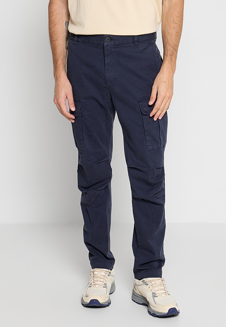 Marinefarbene Cargo-Hosen aus glattem Baumwollstoff mit Seitentaschen und klassischer Passform, kombiniert mit beigen Sportschuhen.