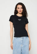 Frau mit lockigem dunklem Haar, die ein schwarzes eng anliegendes T-Shirt mit kurzen Ärmeln und weißem Text sowie hellblaue hoch taillierte Jeans trägt, steht vor weißem Hintergrund.