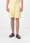Mann in hellgelben Shorts mit Zugband und schwarzen Sandalen, der vor einem weißen Hintergrund steht.