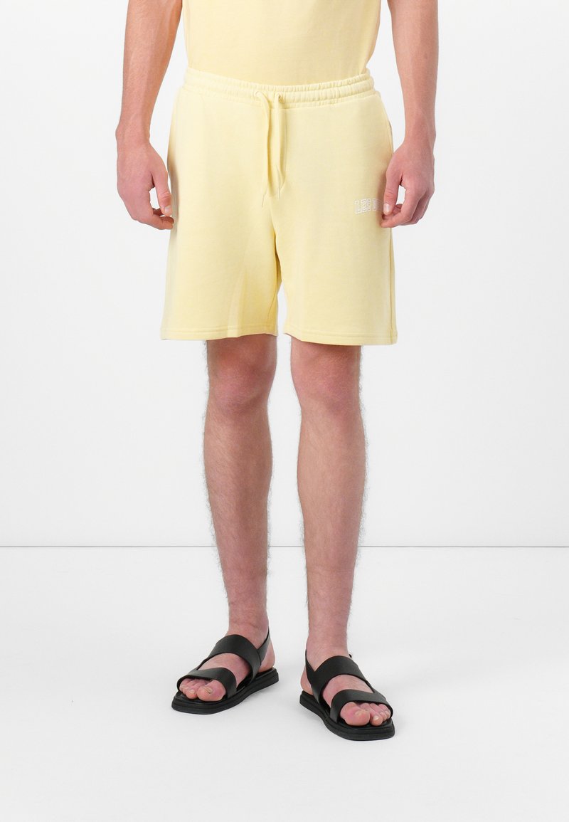 Mann in hellgelben Shorts mit Zugband und schwarzen Sandalen, der vor einem weißen Hintergrund steht.