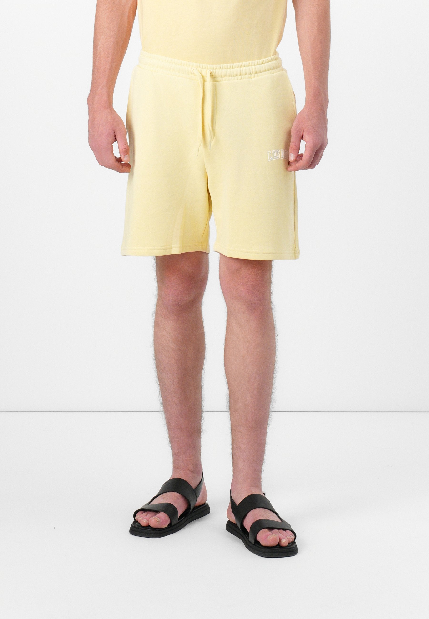 Mann in hellgelben Shorts mit Zugband und schwarzen Sandalen, der vor einem weißen Hintergrund steht.