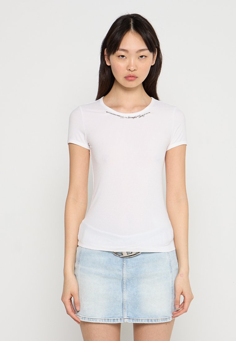 Weißes geripptes T-Shirt mit kurzen Ärmeln und Rundhalsausschnitt, versehen mit dekorativen Stickdetails über der Brust, kombiniert mit einem hellen Jeansrock.