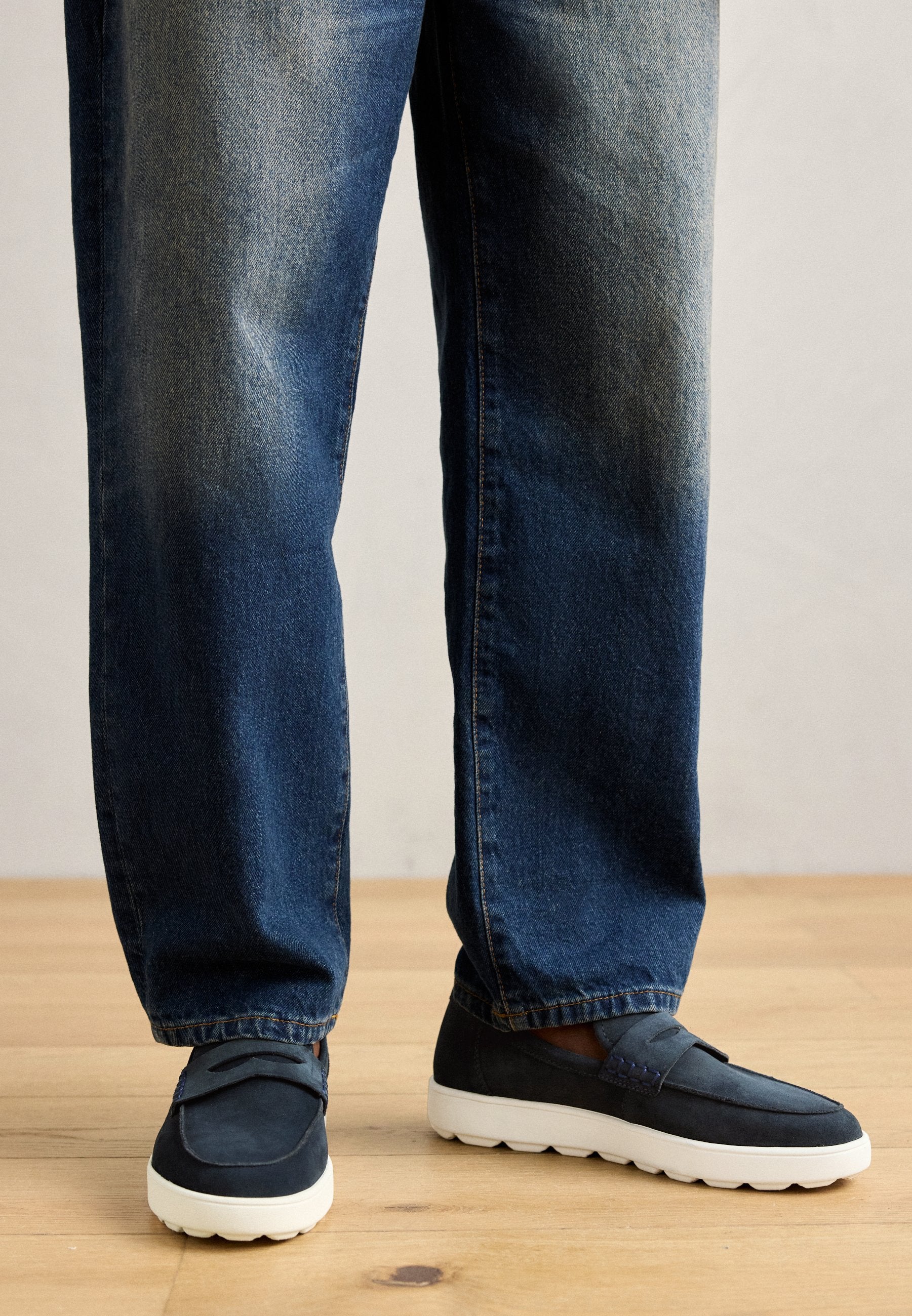 Marineblaue Wildleder-Slipper mit weißer Gummisohle, kombiniert mit verwaschenen blauen Jeans im geraden Schnitt. Die Schuhe verfügen über einen einfachen gestickten Akzent.