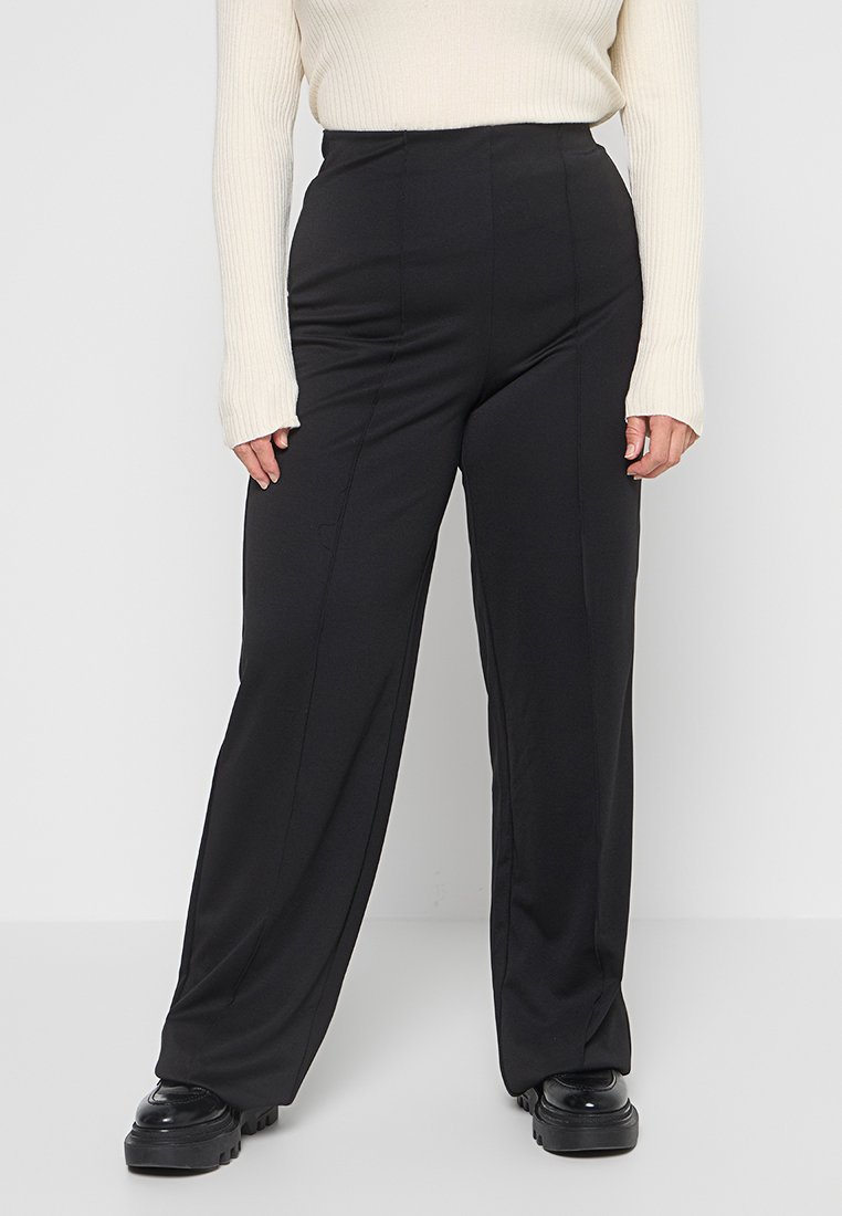 Schwarze weit geschnittene Hose mit hoher Taille und glatter Textur, ausgestattet mit vorderen Nähten und einer geraden Silhouette. Kombiniert mit einem leichten Pullover.