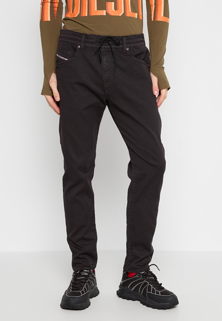 Schwarze Denim-Hosen mit Kordelzugbund, fünf Taschen und dezenten Nähdetails, kombiniert mit schwarzen Schnürschuhen mit roten Akzenten.