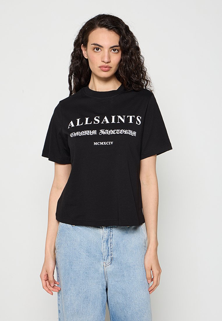 Frau mit lockigem dunklem Haar, die ein schwarzes AllSaints T-Shirt und hellblaue Jeans trägt, steht vor einem schlichten Hintergrund.