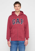 Junger Mann trägt einen weinroten Hoodie mit Reißverschluss und „GAP“-Logo auf der Brust sowie blaue Jeans, steht vor einem schlichten hellen Hintergrund.