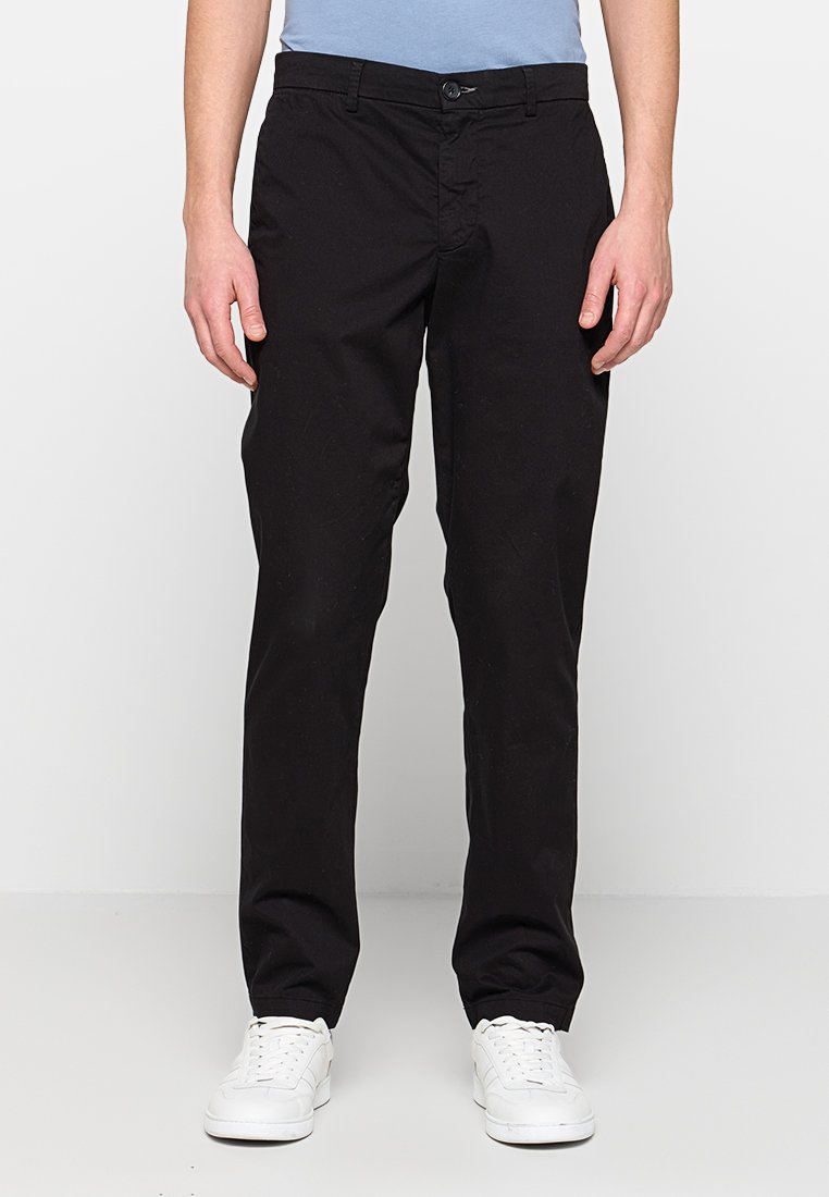Schwarze Baumwollmisch-Hose mit geradem Bein, ausgestattet mit Knopfverschluss und Gürtelschlaufen. Kombiniert mit weißen Turnschuhen.