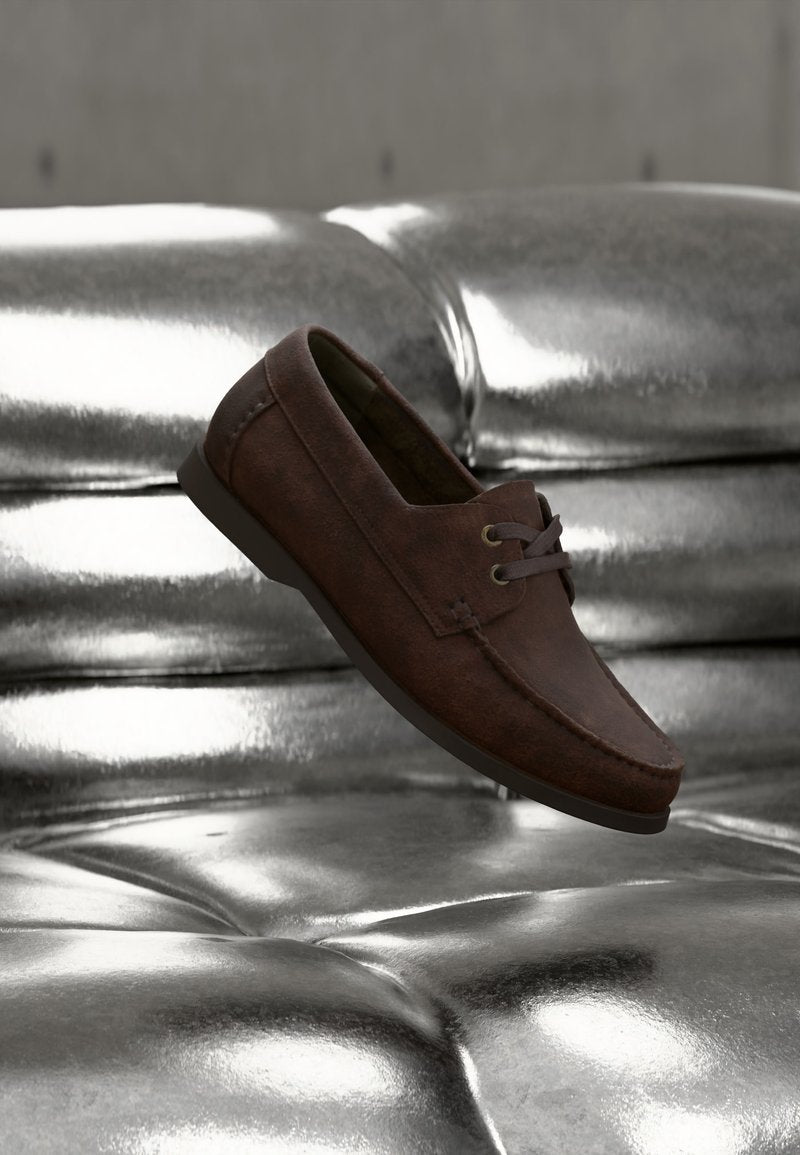 Brauner Lederloafer mit dunklen Schnürsenkeln, der auf glänzendem, gestepptem silberfarbenem Lederpolster ruht.