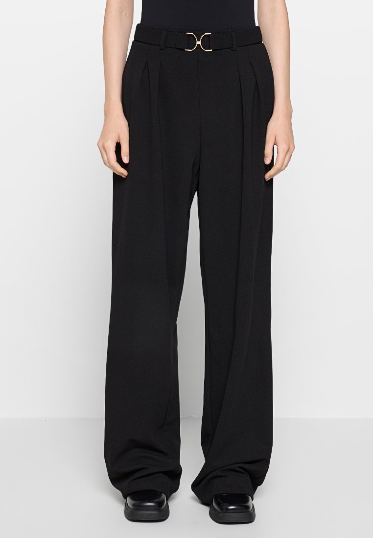 Schwarze weite Hosen mit Falten, einem passenden Gürtel mit einer goldenen runden Schnalle und einer glatten Textur. Kombiniert mit einem enganliegenden Top.