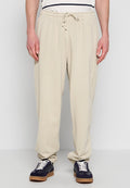 Mann trägt beige Jogginghose mit Kordelzug in der Taille und schwarze Sneakers mit weißen Schnürsenkeln, steht vor hellem Hintergrund.