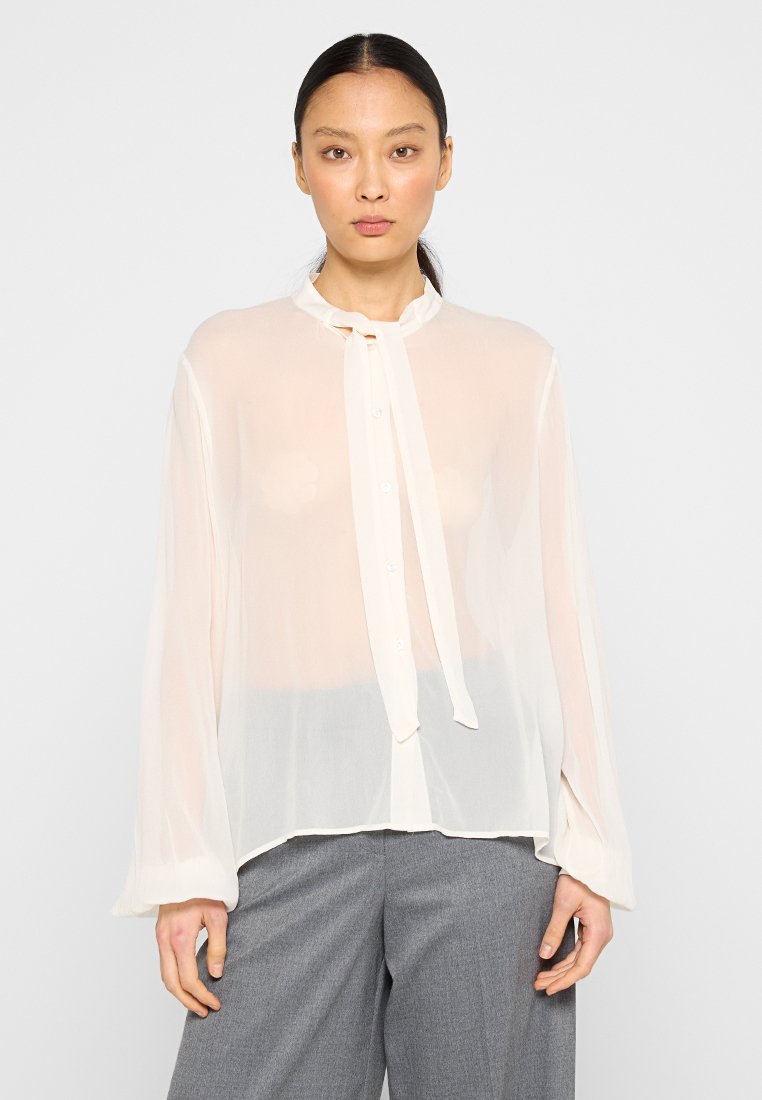Frau mit dunklen Haaren in einem tiefen Pferdeschwanz, die eine transparente weiße Bluse mit Halsbinde und graue Hosen trägt, steht vor einem schlichten hellen Hintergrund.
