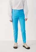 Person, die leuchtend blaue Slim-Fit-Hosen, eine weiße langärmlige Bluse mit Lochmuster und beigefarbene offene Sandalen mit Schnallen trägt, steht auf einem weißen Boden.