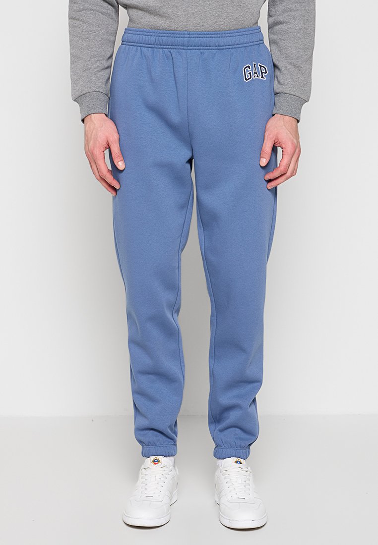 Mann trägt blaue GAP-Jogginghosen mit elastischen Bündchen, ein graues Sweatshirt und weiße Turnschuhe, steht vor einem weißen Hintergrund.