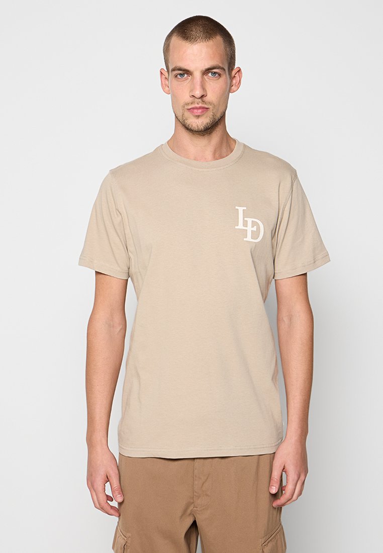 Beige Baumwoll-T-Shirt mit kurzen Ärmeln und Rundhalsausschnitt, mit einem kleinen weißen Logo auf der linken Brust. Glatte Textur.