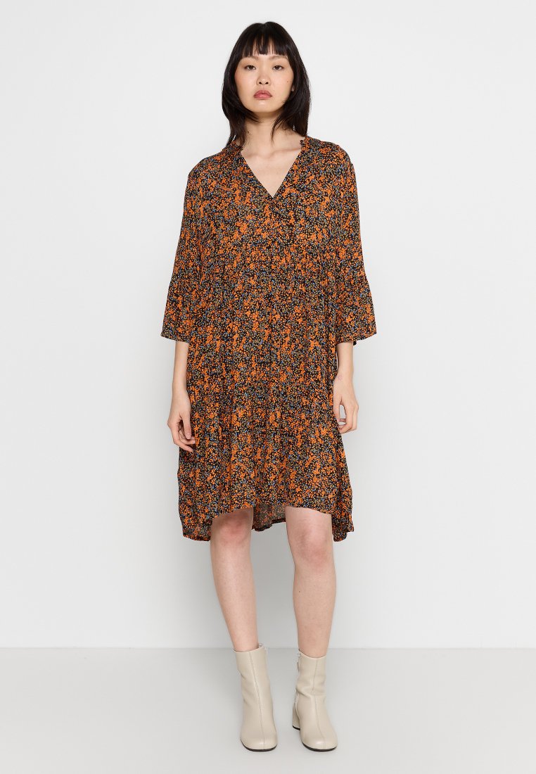 Schwarz-orange-blumiges Kleid mit Dreiviertelärmeln, knielangen Rüschen, V-Ausschnitt und fließender Silhouette. Tragen mit beigen KnöchelStiefeln.
