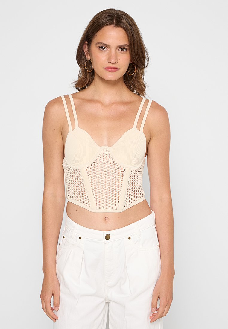 Beige Ripp-Bralette mit transparenter Strickdetails, verstellbaren Trägern und strukturierter Passform, kombiniert mit hochgeschnittenen weißen Hosen.