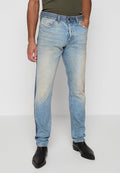 Mann in hellblauen, verwaschenen Jeans, dunkelolivgrünem Hemd und schwarzen, spitzen Stiefeln, der mit einer Hand in der Tasche steht.