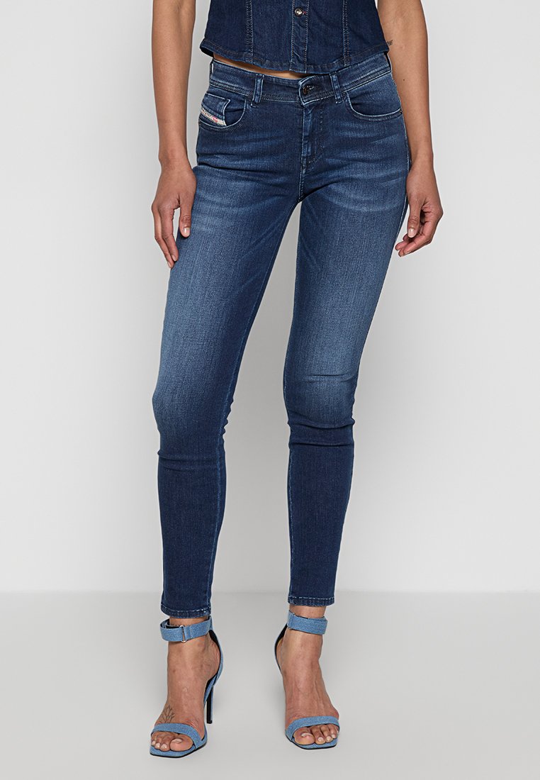 Frau trägt dunkelblaue Skinny-Jeans, ein ärmelloses Denim-Oberteil und hellblaue High-Heel-Sandaletten mit Knöchelriemen und steht vor einem neutralen Hintergrund.