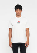 Mann in einem weißen Diesel-T-Shirt mit einem roten und schwarzen Logo auf der Brust und schwarzen Hosen, steht vor einem schlichten weißen Hintergrund.