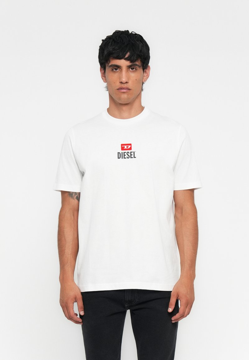 Mann in einem weißen Diesel-T-Shirt mit einem roten und schwarzen Logo auf der Brust und schwarzen Hosen, steht vor einem schlichten weißen Hintergrund.