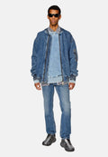 Jeansjacke mit gerippten Bündchen, über einem hellblauen Sweatshirt getragen, kombiniert mit zerrissenen Jeans und schwarzen Sportschuhen. Schwarze Sonnenbrille.