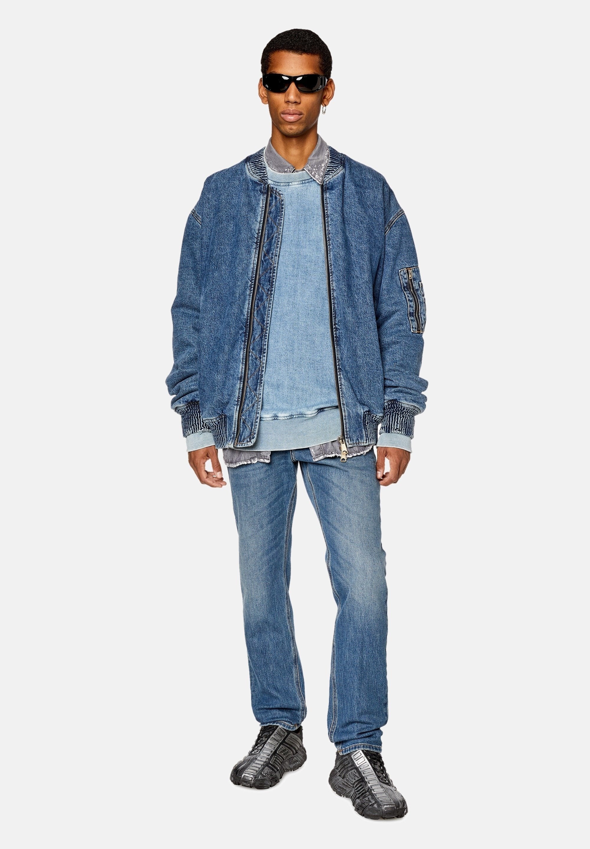 Jeansjacke mit gerippten Bündchen, über einem hellblauen Sweatshirt getragen, kombiniert mit zerrissenen Jeans und schwarzen Sportschuhen. Schwarze Sonnenbrille.