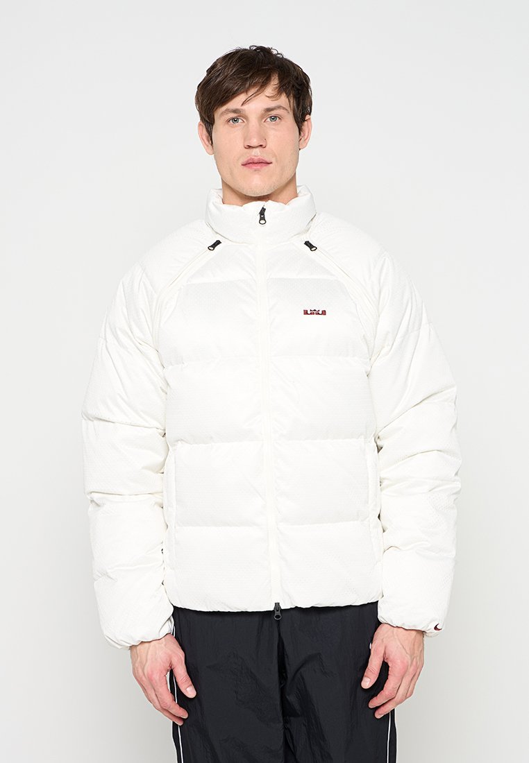 Mann trägt weiße Steppjacke mit Reißverschluss und schwarze Jogginghose, steht vor einem schlichten weißen Hintergrund.