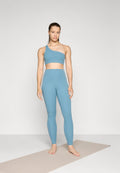 Hellblaues Sportset bestehend aus einem einseitig geschnittenen Crop-Top und hochgeschnittenen Leggings mit einer glatten Textur, auf einer Yogamatte stehend.