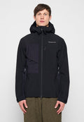 Mann trägt eine schwarze Kapuzenjacke von Peak Performance mit durchgehendem Frontreißverschluss und olivgrüner strukturierter Hose, steht vor einem einfarbigen Hintergrund.