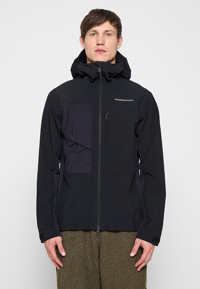 Mann trägt eine schwarze Kapuzenjacke von Peak Performance mit durchgehendem Frontreißverschluss und olivgrüner strukturierter Hose, steht vor einem einfarbigen Hintergrund.