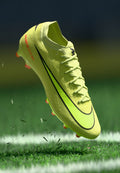 Neon-gelbe Fußballschuhe mit schwarzen Akzenten und strukturiertem Obermaterial, ausgestattet mit Schnürverschluss und orangenen Stollen, über dem Gras erhoben.