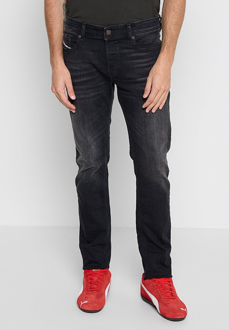 Schwarze Jeans aus Denim mit schlanker Passform, mit subtiler Ausbleichung, fünf Taschen und einem braunen Knopfverschluss. Getragen mit roten Sportschuhen.