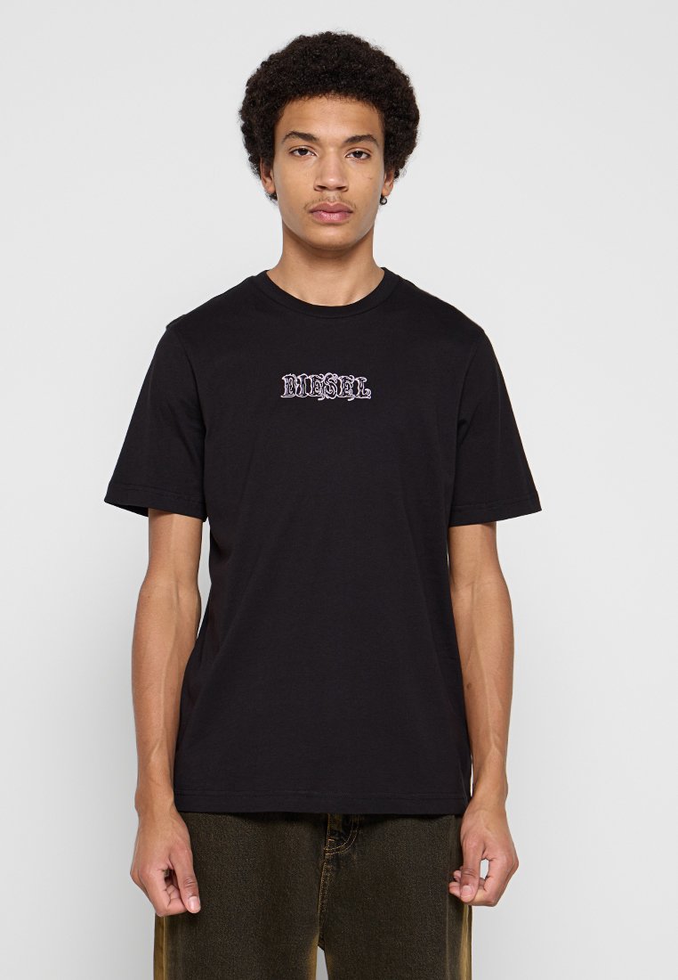 Junger Mann mit kurzen lockigen Haaren, der ein schwarzes Diesel-T-Shirt und dunkle Hosen trägt, vor einem einfarbigen hellen Hintergrund stehend.
