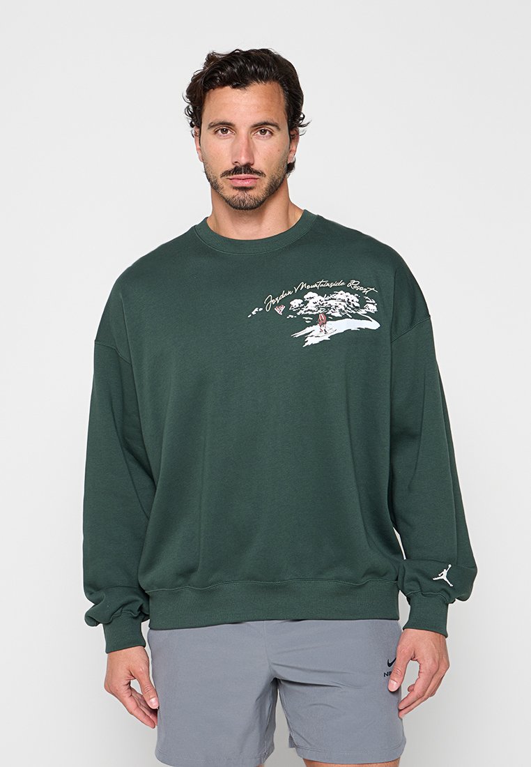 Mann mit kurzen dunklen Haaren und Bart, der einen dunkelgrünen Pullover mit einem Aufdruck eines Bergresorts und graue Nike-Shorts trägt.