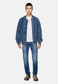 Junger Mann in einer übergroßen Jeansjacke, weißem T-Shirt, verblichenen blauen Jeans, hellbraunen quadratischen Stiefeln und einer Anhänger-Halskette, der vor einem weißen Hintergrund steht.