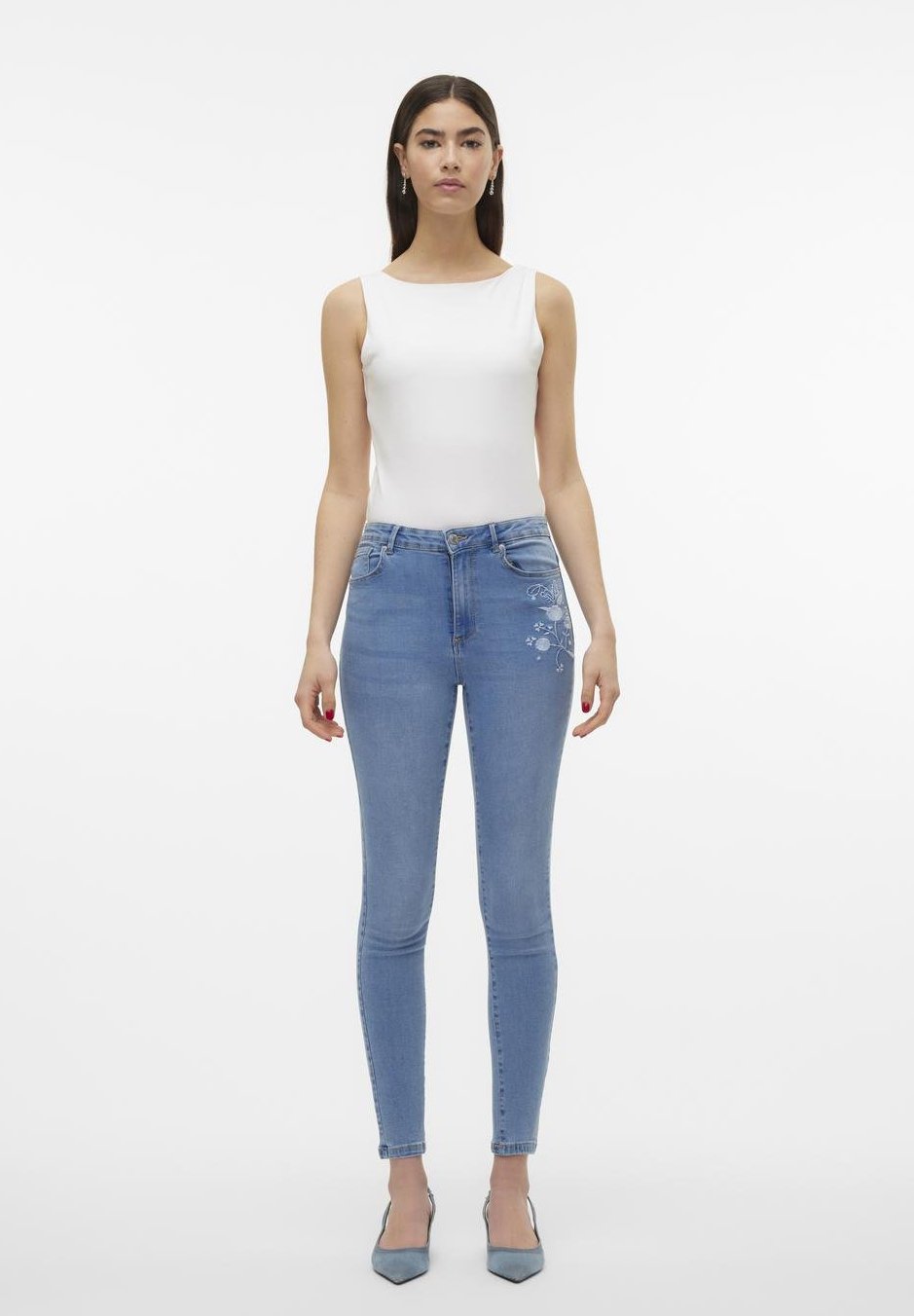 Skinny Jeans aus hellblauem Denim mit floralem Stickdetails am Oberschenkel, getragen mit einem weißen, figurbetonten Tanktop und hellblauen Absatzschuhen.